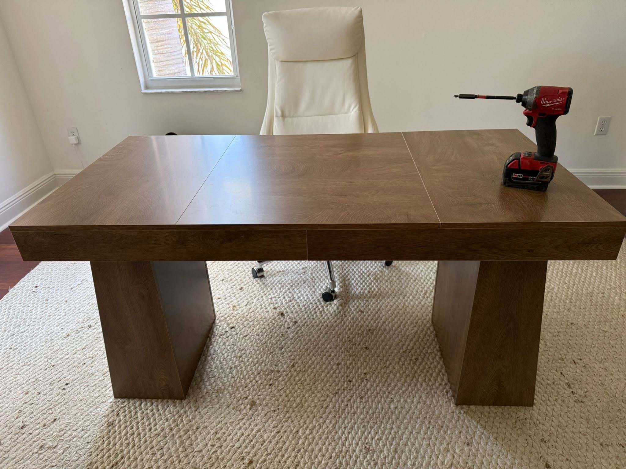 Wooden conference table with extension leaf, cream chair, and Milwaukee power drill on surface